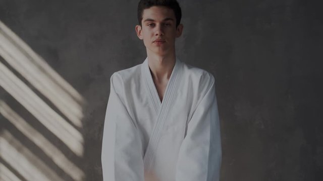 Close up. Handsome guy in white kimono makes a welcome gesture or fight stance. He puts his hands together and looking at the camera. Zoom in. Taekwondo traditional martial arts.