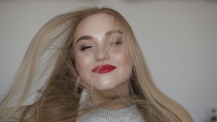 Indoors slow motion closeup face shot of young smiling woman with long blond hair fluttering on the wind, bright red lips, dressed grey sweater, posing against white wall natural light, model looking