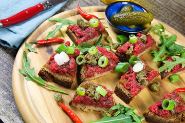 Hearty hefty german snack: isolated wood plate with roasted beef tatar garlic bread, spring onions, capers, pickled cucumbers (gherkins) and rucola salad leaves
