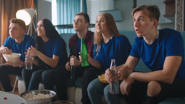 A group of friends in blue t-shirts are emotionally worried watching TV sitting at home on the couch, with popcorn and drinks, and enjoying a goal scored.