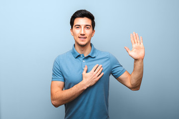 handsome young man with neutral smile in blue polo shirt with hand on chest giving oath against blue background. studio shoot - obrazy, fototapety, plakaty