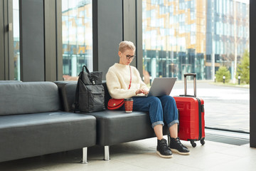 Horizontal full shot of stylish blond woman wearing eyeglasses sitting in departure lounge surfing Internet using her laptop