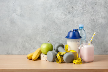 Protein cocktail, dumbbells, measuring tape, water bottle and fresh fruits on wooden table