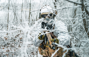 Naklejka premium Airsoft man in white camouflage uniform and machinegun with optical sight. Soldier in the winter forest between branches. Horizontal photo.