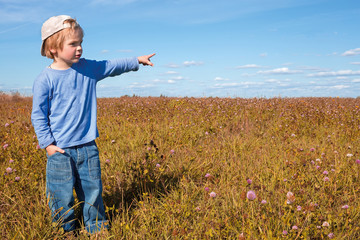 A boy in a blue t-shirt is standing in the field. The boy points a finger at the sky.