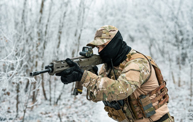 Fototapeta premium Airsoft man in camouflage uniform and machinegun with optical sight. Soldier in the winter forest. Side view.
