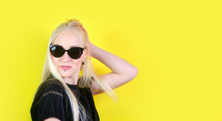 Young cheerful blonde girl in sunglasses rejoices and poses on a yellow studio background.