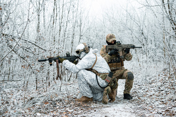 Fototapeta premium Two men in camouflage uniform with machineguns back to back. Soldiers with muchinegun stood on knelt in the winter forest. Side view.