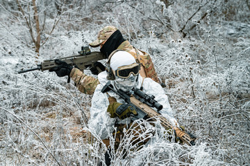 Two men in camouflage white, green uniform with machineguns back to back. Soldiers with muchinegun stood on knelt in the winter grass.