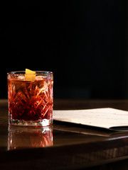 Old fashioned cocktail in glass with dark background on wooden bar desk or table  and a list of paper or menu on it