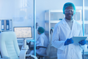 Horizontal medium portrait of handsome young male scientist wearing white lab coat, medical cap and protective eyewear holding tablet PC looking at camera