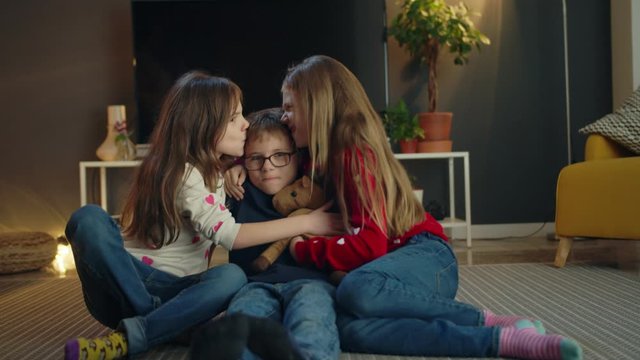 Cheerful Caucasian lovely young kids triplets having fun together in modern living room, attractive little brother and sisters kissing and hugging sitting on floor carpet in casual jeans indoors
