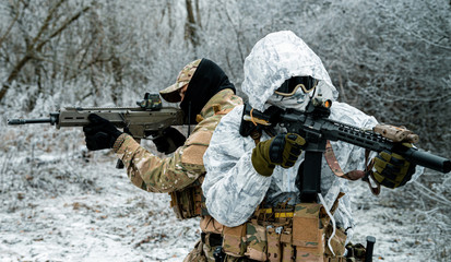 Fototapeta premium Two men in camouflage white, green uniform with machineguns back to back. Soldiers with muchinegun in the winter forest. Horizontal photo, side view.