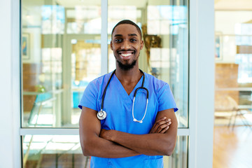 Portrait of a friendly male doctor or nurse wearing blue scrubs uniform and stethoscope, with arms crossed in hospital - obrazy, fototapety, plakaty