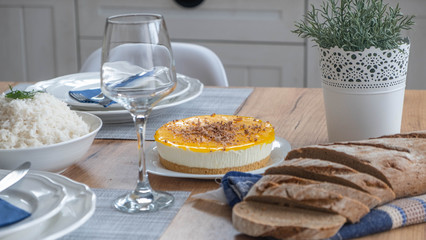 Yellow cheesecake with chocolate on top with brown bread on table with glass and rice bowl in background. 