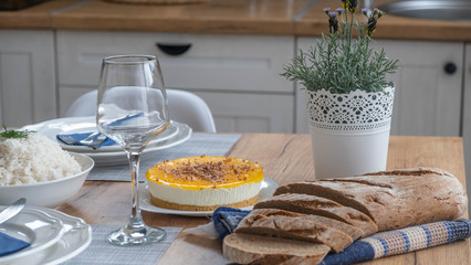 Yellow cheesecake with chocolate on top with brown bread on table with glass and rice bowl in background. 