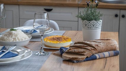 Yellow cheesecake with chocolate on top with brown bread on table with glass and rice bowl in background. 