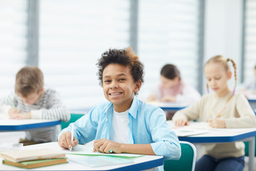 Horizontal medium close up portrait of happy mixed-race boy with kinky hair sitting at school desk...