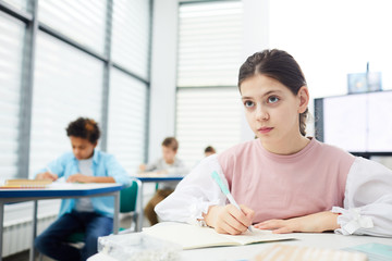 Pensive twelve-year-old schoolgirl with dark hair wearing pink and white outfit looking at blackboard and making notes, copy space