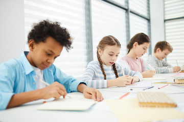 Group of four middle school students sitting together at desk in modern classrooms completing task in their notebooks, copy space