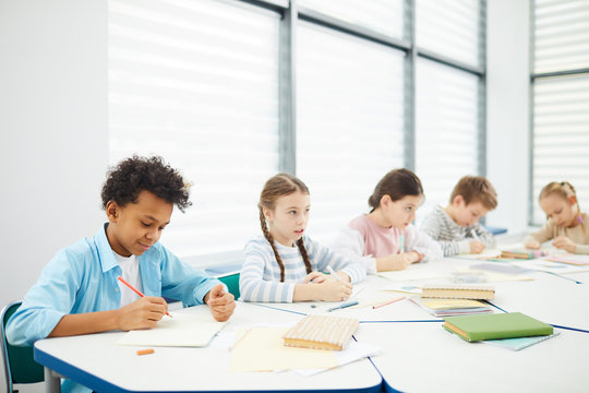 Group Of Five Middle School Students Wearing Casual Outfits Sitting At Desk In Modern Classroom Completing Task, Horizontal Shot, Copy Space