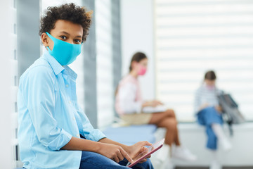Horizontal portrait of unrecognizable Black boy wearing mask sitting in queque to school medical...