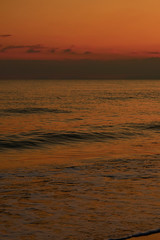 Colorful sunset at the tropical beach, sun behind clouds reflects on water and waves with foam hitting sand.