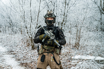 Fototapeta premium Airsoft man in black camouflage uniform and short machinegun with optical sight. Soldier in the winter forest. Horizontal photo front view.