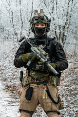 Fototapeta premium Airsoft man in black camouflage uniform and short machinegun with optical sight. Soldier in the winter forest. Vertical photo front view.