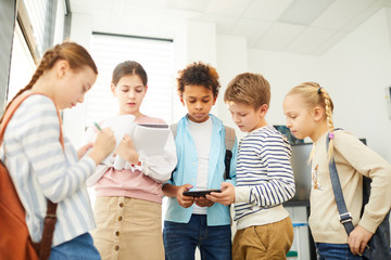 Group of five middle schoolers standing together in school corridor searching information about...