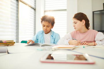 Fototapeta na wymiar African American boy and Caucasian girl sitting together at school desk in modern classroom, girl looking at boy's textbook, copy space