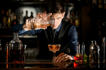 Young bartender gently pours brown cocktail into glass.
