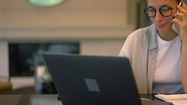 Caucasian female economist sitting at desktop with modern netbook for digital marketing and making mobile conversation for online communication with colleague, young woman in spectacles calling via ap