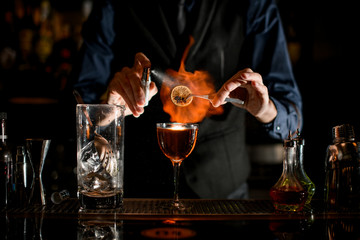 Professional man at the bar sprinkles burning slice of citrus for cocktail.
