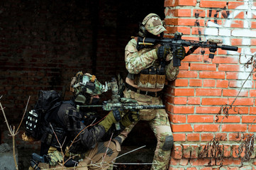 Two men in camouflage cloth and black uniform with machineguns. Soldiers with muchinegun aims aiming standing beside wall.