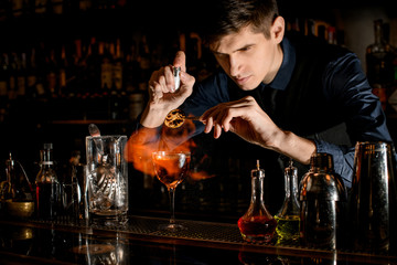 Young bartender at the bar sprinkles burning slice of citrus for cocktail.