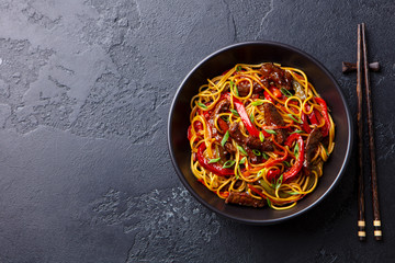 Stir fry noodles with vegetables and beef in black bowl. Slate background. Copy space. Top view.