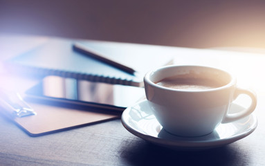Close-up of white coffee cup with mobile phone and notebook on wooden desk with vintage light flare,