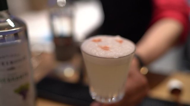 bartender's hand offering a Pisco Sour, the iconic Peruvian cocktail, inside a bar. Captured in slow motion, the scene emphasizes the drink's frothy top,  the art of mixology.