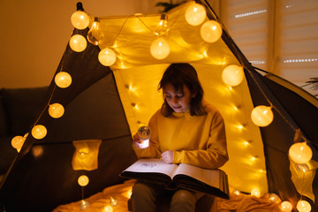 Young female child reading encyclopedia in a home made livingroom tent with light balls.