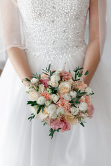 Bridal bouquet of white roses and peach peonies with pink eustoma and white chrysanthemum, decorated with green eucalyptus leaves, held by a bride in white wedding dress with decorated bodice