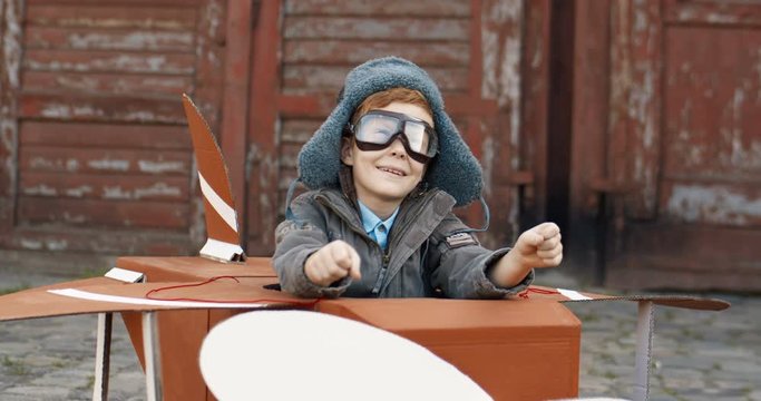 Happy cheerful small Caucasian boy with red hair in hat and glasses sitting outdoors in wooden toy model of airplane and playing like rulling. Cute kid dreaming to be aviator when grow up.