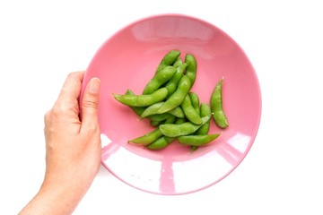 Top eye view pieces of girl hand are holding many green Edamame soybeans Japan in circle pastel pink color plate isolated on vintage white table background at a kitchen room, ready for serving.