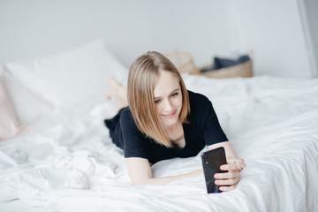 Young beautiful woman lies in black sweater on bed and uses smart phone for online chatting work, smiling