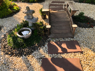 Japanese style garden consisting of a small wooden bridge across the white gravel floor, Decorated with stone lanterns and small trees.