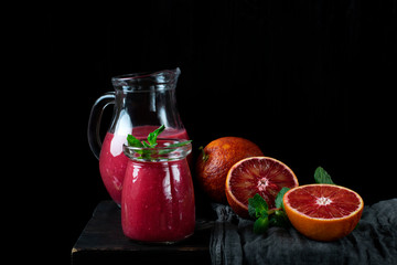 Smoothie with blood orange juice in the glass jar and jug on the edge of the wooden table. Still life against the black background