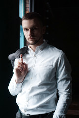 View in profile of a young businessman, dressed in a white shirt standing near the window on a dark walls and looking at the camera. Vertical photo