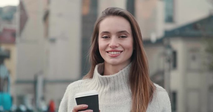 Portrait of Beautiful female Model posing on Camera and walking in front of Objective. Wearing Casual stylish Sweater. Having nude makeup and Healthy brunette Hair. Holding hot Coffee in Paper cup.