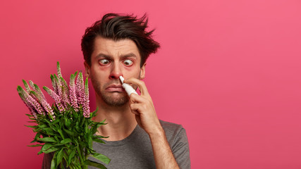 Sick man poses with allergy trigger, uses nasal drops to cure runny red sore nose, has redness...