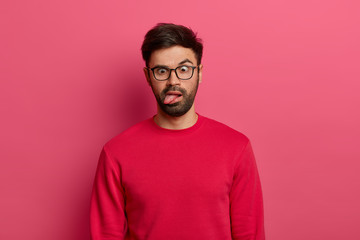 Photo of crazy bearded man sticks out tongue, crosses eyes, feels tired and bored, wears glasses...